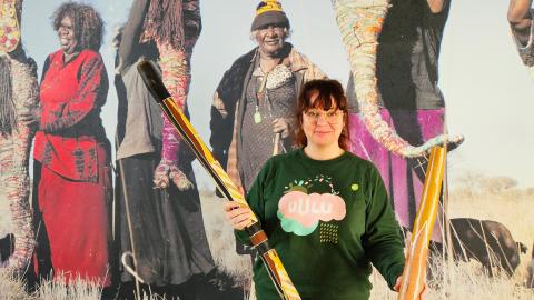 Nainen pitelee käsissään kahta isoa puista soitinta, taustalla aboriginaalitaiteilijoita esittävä kuva.