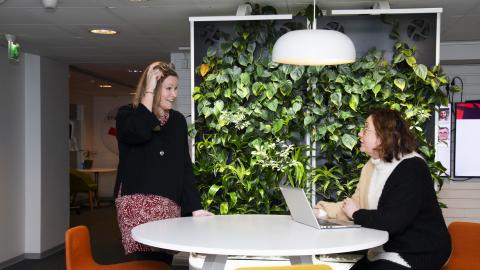 Kaksi henkilöä juttelee pyöreän pöydän ääressä. Toinen seisoo ja toinen istuu koneen ääressä. Takana on viherseinä.