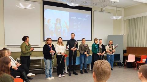 Sertifioitujen kukittamistilaisuudessa otetun kuvan keskellä sertifioidut ihmiset rivissä, yleisön ympäröimänä.