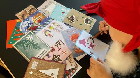 Lapsi tonttulakissa piirtämässä joulukorttia. Pöydällä kortteja.