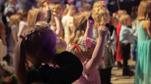 Juhla-asuinen lapsi katselee edessään olevia muita lapsia, jotka tanssivat. 