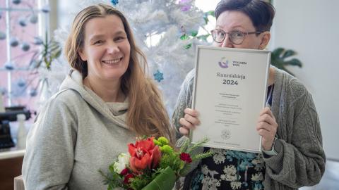 Kaksi henkilöä, joista toinen pitää käsissään kukkakimppua ja toinen kunniakirjaa