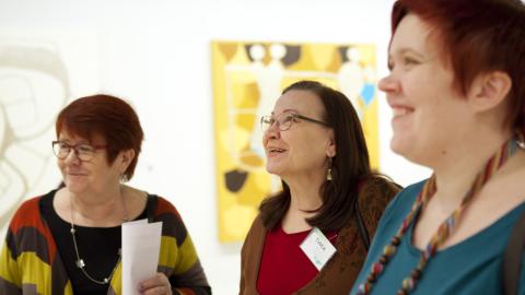 Kolme naista katsoo taidetta yhdessä, taustalla näkyy keltainen maalaus.