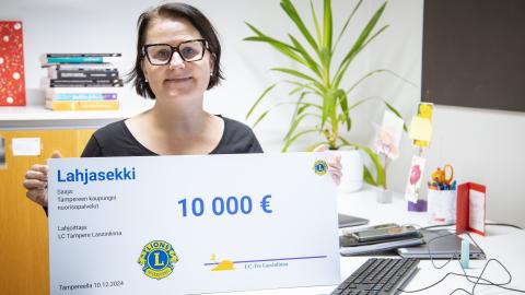 Nainen seisoo pöytänsä ääressä iso 10 000 lahjasekki kädessään ja hymyilee