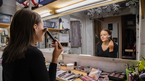 Nuori meikkaa itseään paksulla siveltimellä ison peilin edessä. Peilin alla olevalla pöydällä on erilaisia meikkejä.