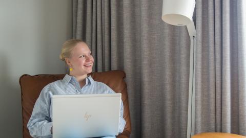 Vaaleatukkainen henkilön istuu tuolilla ja katsoo iloisesti sivulle. Taustalla näkyy harmaat verhot ja lattialamppu.