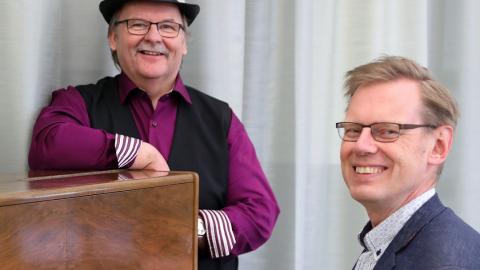 Sinitakkinen mies istuu pianon ääressä, toinen violettipaitainen mies nojaa pianoon taaempana. Molemmat hymyilevät.