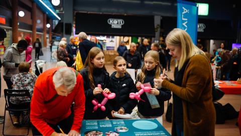 People of different ages at the Tampere Residents’ Evening in a shopping center. At the table, there are two adults writing on paper and three young people with balloons.