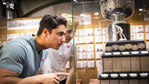 Tummatukkainen mies osoittaa vitriinissä olevaa suurta jääkiekkopokaalia, vaaleahiuksinen mies taustalla.