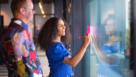 Kaksi henkilöä sivuttain toimiston aulassa, jossa isot ikkunat. Etumainnen kirjoittaa ikkunassa kiinni olevaan muistilappuun ja hymyilee. Taaempana oleva henkilö katselee ja hymyilee hänkin. 