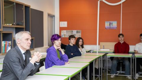 Ministeri ja opiskelijat istuvat rinnakkain luokkatilassa, jonka yksi seinä on voimakkaan oranssin värinen.