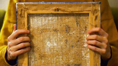 Keltapaitainen henkilö pitää sylissään vanhaa taulun kehystä, josta näkyy teoksen tausta.