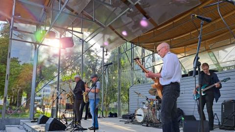 A jazz band of middle-aged men performs on an outdoor stage in sunny weather.