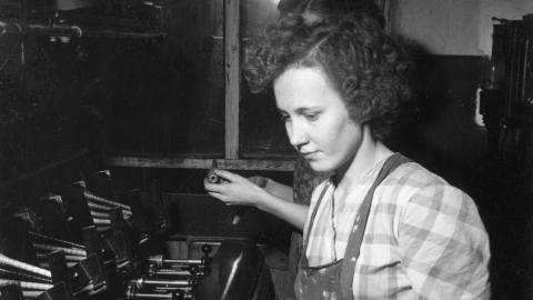 Mustavalkoinen kuva Finlaysonin puuvillatehtaan kutojasta 1930-luvulla. Ruutupaitainen nainen, jolla on käsissään laatikollinen lankapuolia.