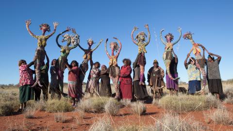 Australian aavikolla kuvattuna joukko aboriginaaleja pitelemässä ilmassa luonnonmateriaaleista valmistettuja nukkeja. Taivas taustalle on sininen.