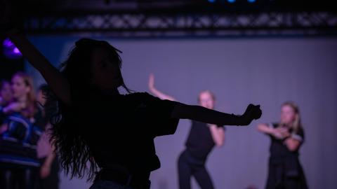 Pitkähiuksinen henkilö tanssii kuvan etualalla. Taustalla epätarkkana muita tanssijoita.