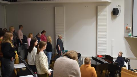 Henkilöt seisovat penkkiriveillä ja venyttelevät. Flyygelin vieressä istuu kuoronjohtaja.