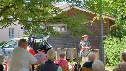Nainen soittaa ukulelea kesäisessä puutarhassa ja tuoleilla istuvat istuvat ikäihmiset laulavat mukana.