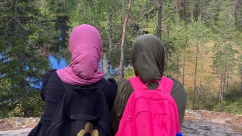 Kaksi nuorta henkilöä istuu vierekkäin korkealla kalliolla ja katselee metsäistä maisemaa, jossa näkyy myös pieni lampi. 