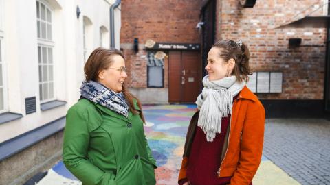 Kaksi hymyilevää naista katsoo toisiaan. Toisella on vihreä ja toisella oranssi takki.