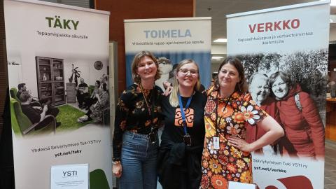 Kolme naista seisoo hymyillen esittelytiskin takana messutilaisuudessa.