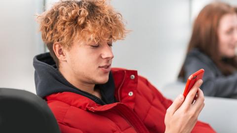 A young person is sitting and looking at their smartphone, which they are holding in their hand. Another person in the background.