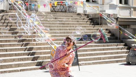 Punaisiin pukeutunut nainen pitää käsissään värikästä harjanvartta ja ihailee saippuakuplia.