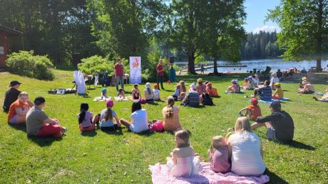 Eri ikäisiä ihmisiä istumassa nurmikolla järven rannalla