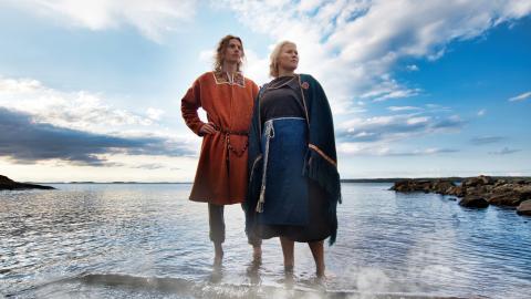 Mies ja nainen seisovat matalassa vedässä rannalla, taustalla sinistä taivasta ja pilviä. Vedästä nousee höyryä.