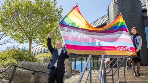 Two people wave a large 13-stripe pride flag of Tampere&#039;s own on the steps of the Metso Main Library.