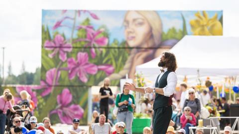 Mies esiintyy joukolle ihmisiä kesäisenä päivänä. Taustalla värikäs seinämalaus.