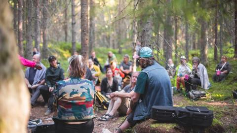 A large group of people are sitting in the Kintulammi forest listening to a musical performance.
