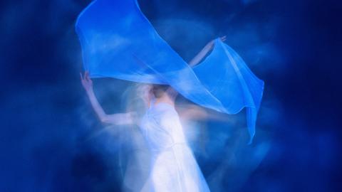 A person in a white dress dances with a flowing blue fabric against a dark blue background.