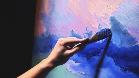 Käsi pitelee pensseliä, joka maalaa sinisiä ja vaaleanpunaisia sävyjä kankaalle.