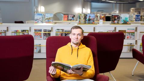Henkilö istuu nojatuolissa pääkirjasto Metson lehtisalissa ja pitää käsissään avointa lehteä. Taustalla on salin lehtikalusteita. 