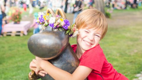 Punapaitainen poika halaa Muumipeikko-patsasta, jolla on kukkaseppele päässä. Taustalla ihmisiä kesäisessä puistossa.