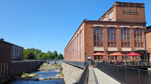 Vapriikin punatiilinen rakennus ulkoa kuvattuna. Edustalla ravintolan ulkoterassi, jossa varjoja ja ruokailijoita. Rakennuksen reunassa alapuolella virtaa koski.