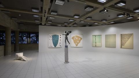 Vaaleaseinäinen näyttelytila, jossa keskellä harmaa patsas. Taustalla näkyy suuria haaleasävyisiä maalauksia.
