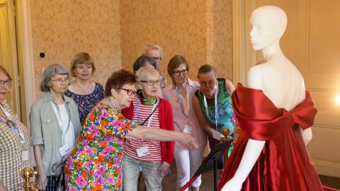 Naiset seisovat museonäyttelyssä ja katselevat punaista, olkaimetonta silkkisatiinista valmistettua iltapukua.