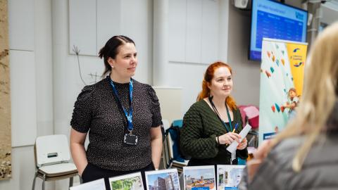 Two experts from the City of Tampere discuss with people at a residents&#039; evening. There are several display stands on the table.