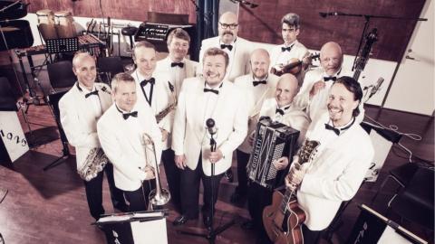 Kymmenhenkinen viihdeorkesteri poseeraa valkoisissa smokkitakeissa soittiminen kanssa.