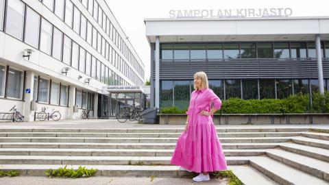 Henkilö seisoo vaaleanpunaisessa pitkässä mekossa rakennuksen edessä, jossa lukee Sampolan kirjasto.