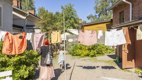 Pyykkejä roikkuu narulla Amurin museokorttelin pihalla kesäpäivänä. Aikuinen ja lapsi kulkevat pyykkien alta.