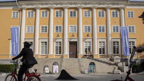 Keltainen suuri rakennus, johon johtaa leveä portaikko. Edessä kaksi pyöräilijää.