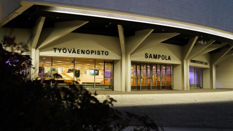 Sampola-rakennus pimeällä. Seinä on valaistu. Etualalla on pensas.