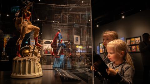 Hämärässä näyttelytilassa kaksi lasta katsoo vitriinissä olevaa Teräsmies ja Ihmenainen figuureja. 