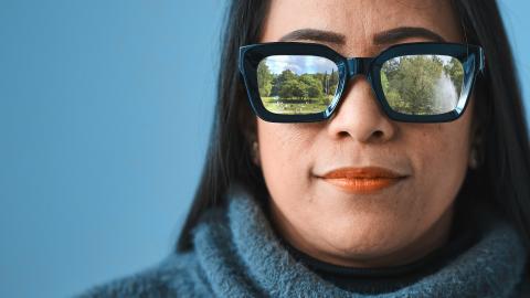 A dark-haired person is looking directly at the viewer. She is wearing thick-framed sunglasses that reflect the image of the park.