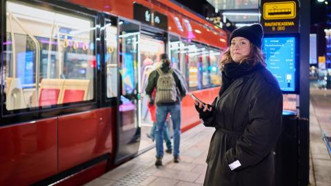 A person is standing at a tram stop holding a smartphone. The tram has stopped and passengers are boarding.
