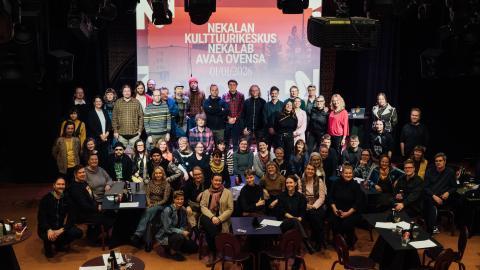 Suuri ryhmä ihmisiä poseeraa yhteiskuvassa. Taustalla valotaulu, jossa lukee &quot;Nekalan kulttuurikeskus Nekalab avaa ovensa 1.1.2026&quot;.
