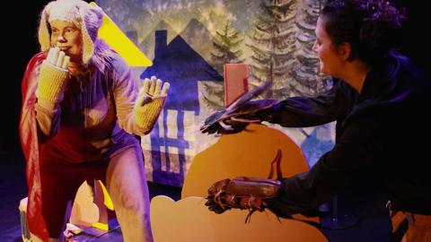 A person dressed in a dog costume holds &quot;paw&quot; in front of their mouth and smiles. In the background are spruce trees and a silhouette of a house.